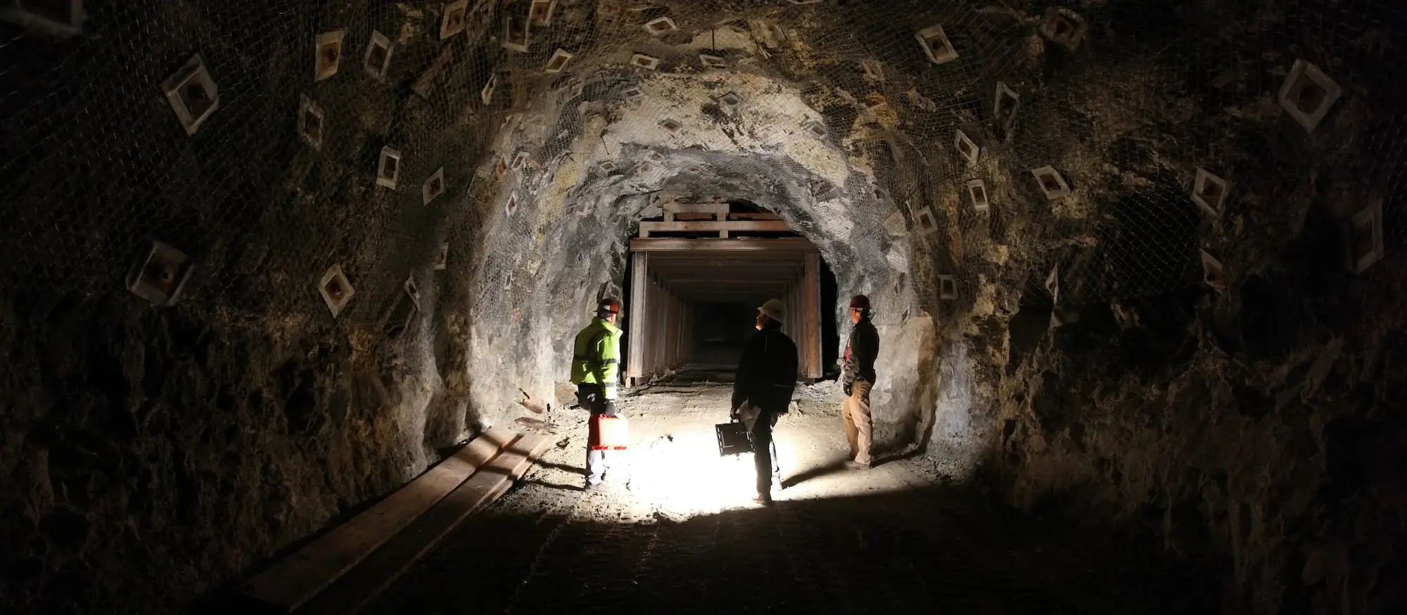 Three people in high-visibility gear standing in a timbered underground drift, lit by a single lamp