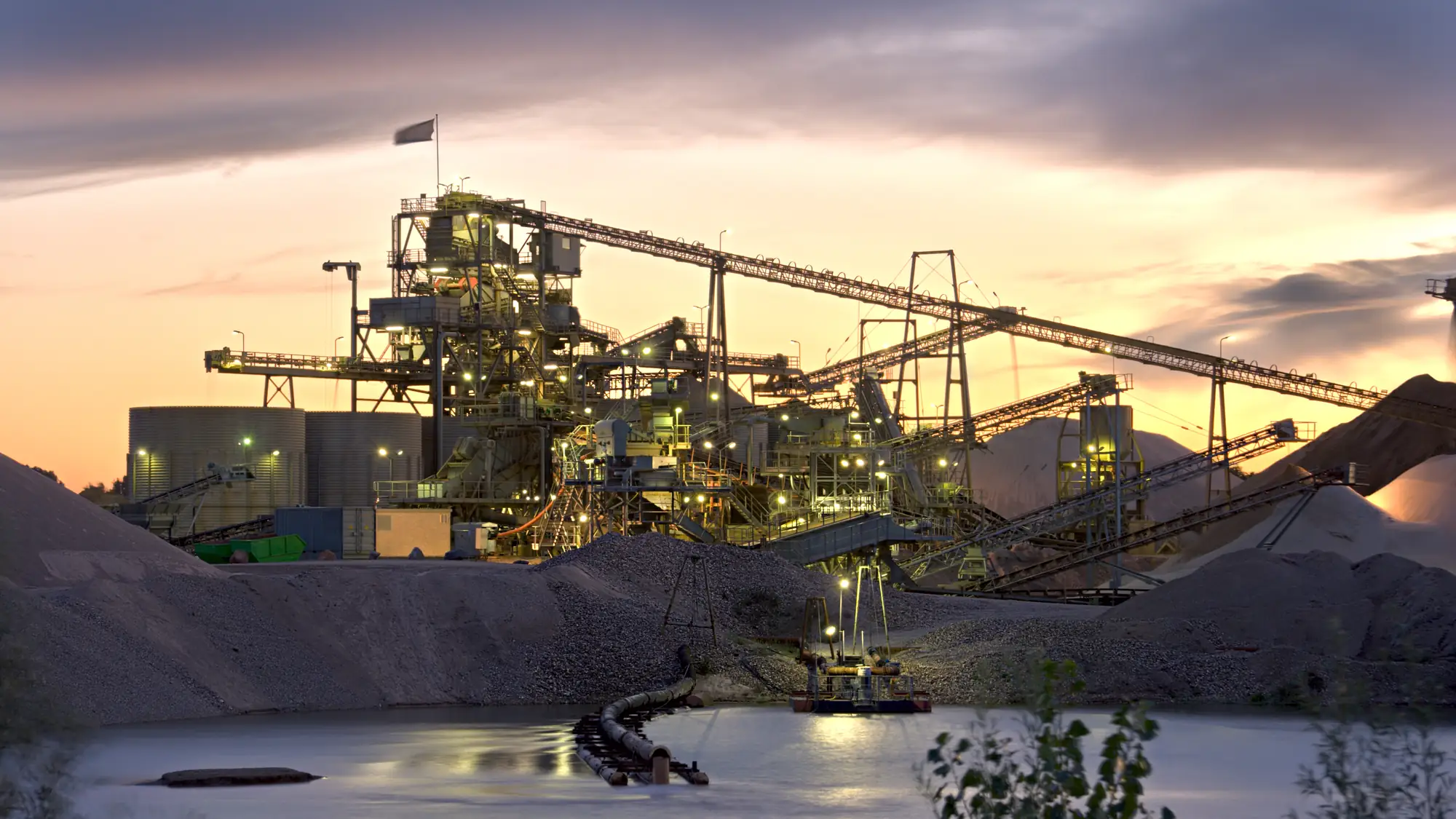 A mineral processing plant lit up at dusk, conveyors and silos above a settling pond