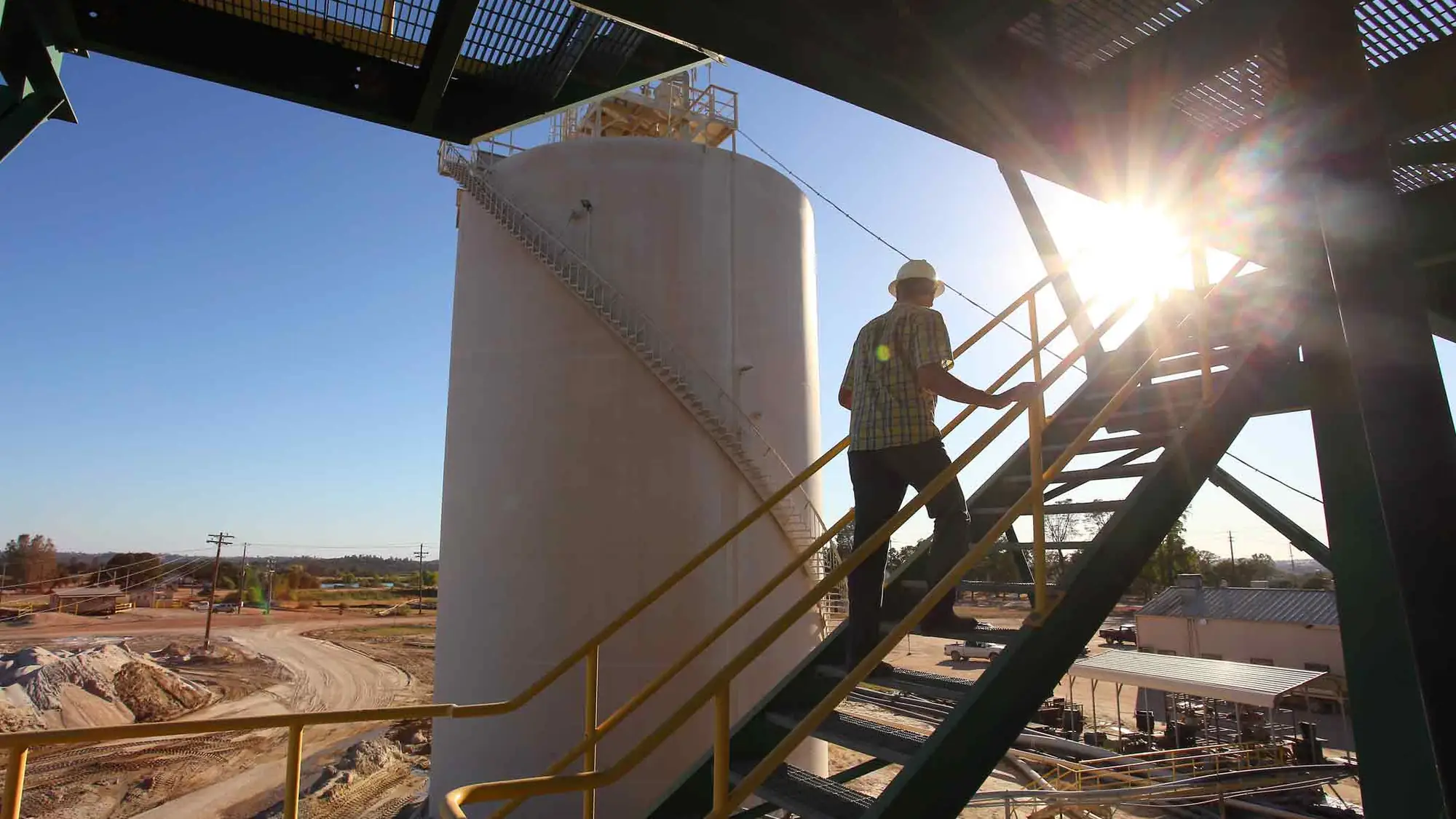 A worker climbing an external stair at a processing plant, sun flaring behind the structure
